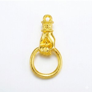 Puede incluir: Un colgante de metal dorado que muestra una mano esculpida agarrando un anillo circular. La mano est&aacute; unida a una pieza superior cil&iacute;ndrica con un peque&ntilde;o lazo para colgar. Toda la pieza tiene un acabado liso y un tono dorado uniforme.