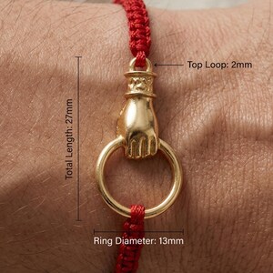May include: A gold-colored bracelet charm featuring a hand holding a ring. The ring has a diameter of 0.5 inches. The total length of the charm is 1.06 inches. The charm is attached to a red woven cord.