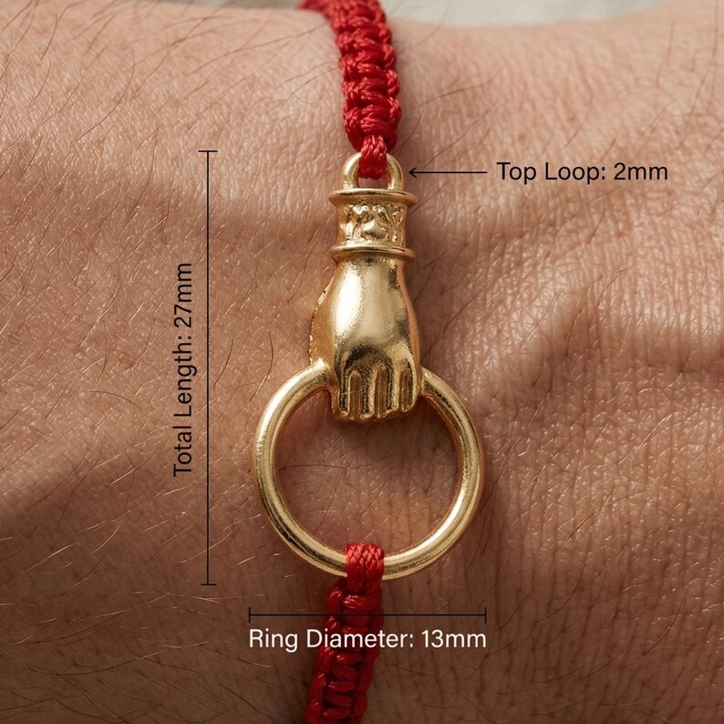 May include: A gold-colored bracelet charm featuring a hand holding a ring. The ring has a diameter of 0.5 inches. The total length of the charm is 1.06 inches. The charm is attached to a red woven cord.