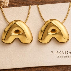 May include: Two gold-toned letter A pendants with a smooth, rounded, and polished finish. The pendants are displayed on a white card with the text 2 PENDANTS and Chain not included printed in the bottom right corner.