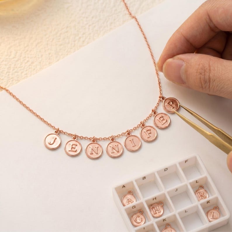 May include: Rose gold necklace with circular pendants spelling "JENNIFER". The necklace is being assembled with tweezers, with individual letter charms in a tray. The pendants are approximately 1.3 cm in diameter.