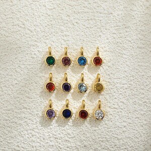 May include: A collection of twelve gold-colored pendants, each featuring a different colored gemstone. The gemstones include green, purple, blue, red, and yellow, set in a round bezel. The pendants are displayed on a textured, off-white surface.