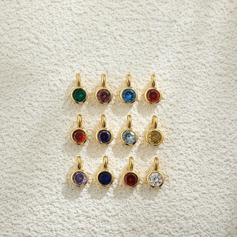 May include: A collection of twelve gold-colored pendants, each featuring a different colored gemstone. The gemstones include green, purple, blue, red, and yellow, set in a round bezel. The pendants are displayed on a textured, off-white surface.