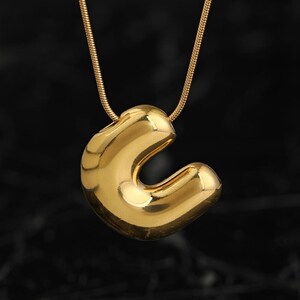 May include: A gold-toned necklace featuring a polished, puffed letter C pendant suspended from a thin, flexible snake chain. The pendant has a smooth, reflective surface with rounded edges, creating a three-dimensional appearance.