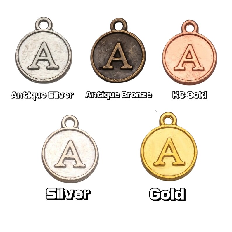 May include: Five metal letter "A" charms in different finishes: antique silver, antique bronze, KC gold, silver, and gold. Each charm is round with a loop for attaching to jewellery or crafts. The names of the finishes are printed below each charm.