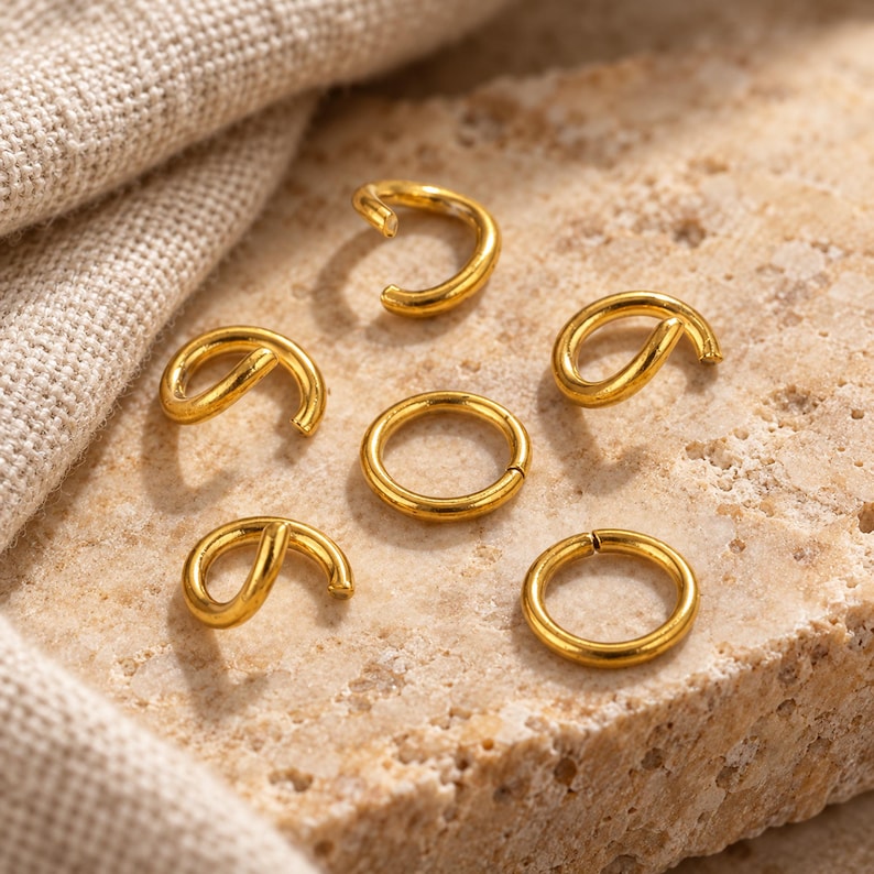 Gold-tone stainless steel open jump rings. Textured hero view. 5mm diameter; 0.8mm wire. Buyer pack: 100 pieces.