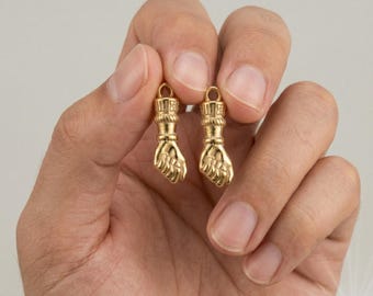 May include: Two gold-colored fist-shaped charms. Each charm has a loop at the top for hanging and detailed hand features. The charms are held in a person's hand against a soft background.