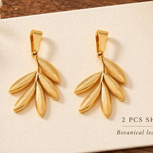 May include: Two gold-toned metal charms shaped like botanical leaf clusters, each featuring a smooth, polished finish and a top bail for attachment. The text 2 PCS SHOWN and Botanical leaf charms is printed on the white surface below the charms.