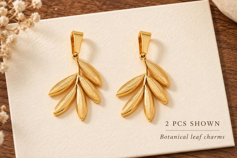 May include: Two gold-toned metal charms shaped like botanical leaf clusters, each featuring a smooth, polished finish and a top bail for attachment. The text 2 PCS SHOWN and Botanical leaf charms is printed on the white surface below the charms.