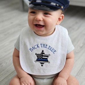 May include: A white baby bib featuring the embroidered text BACK THE BLUE above a black and white star-shaped police badge with a thin blue line. The bib is paired with a dark navy blue police cap that has a silver badge on the front.