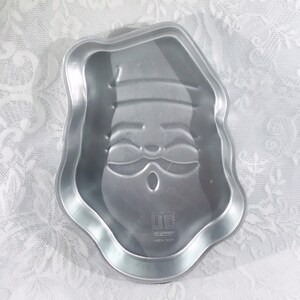 May include: A silver-colored Santa Claus-shaped cake pan. The pan has a detailed face design with a hat, eyes, and a mustache. The pan is made of metal and has a scalloped edge.