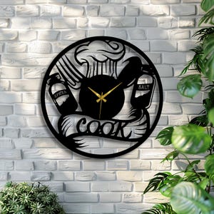Könnte beinhalten: Schwarze Metall-Wanduhr mit kulinarischem Thema. Die Uhr zeigt eine Kochmütze, Utensilien und Gewürzgläser mit dem Wort "COOK" in einem Banner. Die Zeiger der Uhr sind goldfarben, und der Hintergrund ist eine weiße Backsteinmauer.