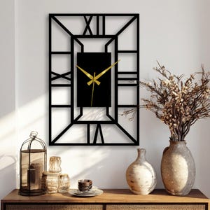 Peut inclure: Horloge murale rectangulaire noire avec des aiguilles dorées et des chiffres romains. L'horloge a un design géométrique et est fixée sur un mur blanc. L'horloge fait partie d'un arrangement de décoration intérieure.