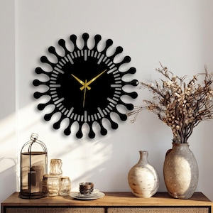 May include: A black wall clock with a unique, stylized design. The clock features a circular shape with a series of rounded protrusions around the edge. The clock hands are gold. The clock is mounted on a white wall, with decorative vases and a candle holder on a wooden surface.