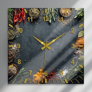 Könnte beinhalten: Eine quadratische Wanduhr mit einem dunklen, strukturierten Hintergrund, der mit verschiedenen Gewürzen, Kräutern und Chilischoten am Rand verziert ist. Das Zifferblatt zeigt goldfarbene Ziffern und passende goldfarbene Uhrzeiger.