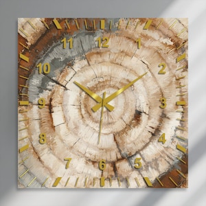 May include: A square wall clock featuring a printed design of concentric tree rings in shades of brown, beige, and grey. The clock face includes gold-coloured hour markers, gold-coloured numerals from 1 to 12, and gold-coloured hour and minute hands.