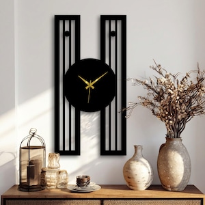 May include: A black wall clock with gold hands, comprising a circular clock face and two vertical striped panels. The clock is mounted on a white wall. Decorative items, including vases, are on a wooden surface below.