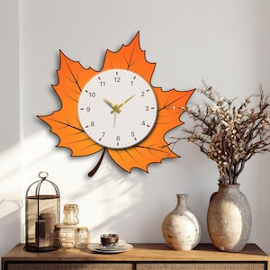 Puede incluir: Un reloj de pared con forma de gran hoja de arce naranja y una esfera circular blanca. El reloj presenta números negros del 1 al 12, finas marcas negras para los minutos y manecillas de color dorado para las horas y los minutos.