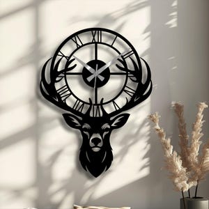 Puede incluir: Reloj de pared de metal negro con diseño de cabeza de ciervo y cornamenta. La esfera del reloj tiene números romanos y agujas plateadas. El reloj está montado en una pared clara.