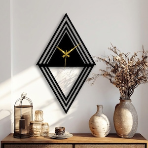 May include: Black and silver geometric wall clock with gold hands. The clock is diamond-shaped with a black center and silver stripes. The clock is mounted on a white wall above a wooden table with decorative items.