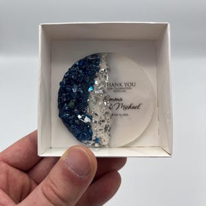 Puede incluir: Un posavasos redondo y decorativo con un semicírculo de cristales azul oscuro y purpurina plateada, y un semicírculo blanco. El texto dice "THANK YOU for celebrating with us! Emma & Michael June 16 2026". El posavasos está en una caja blanca.