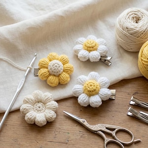 Può includere: Fermagli per capelli a forma di fiore all'uncinetto, gialli e bianchi, con clip in metallo argentato. L'immagine mostra anche un uncinetto, forbici e gomitoli di lana, suggerendo un tema artigianale. I fermagli sono disposti su una superficie di legno.