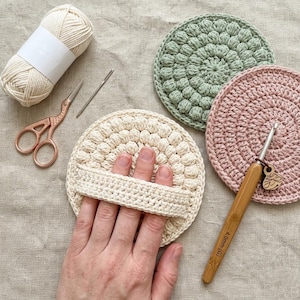 May include: A collection of handmade crochet items. Includes a cream-colored round scrubby with a handle, a green and a pink round scrubby, a ball of yarn, scissors, a needle, and a crochet hook. The crochet hook is 4.0mm.