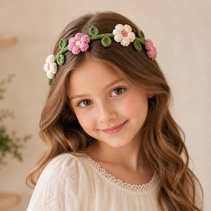 May include: A crochet floral headband featuring pink and white flowers with green leaves. The headband is worn by a person with long brown hair and a cream-colored top. The flowers are evenly spaced along the green band.