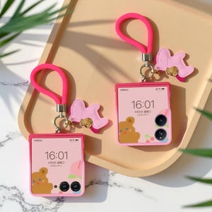 May include: Two pink foldable phone cases with matching wrist straps and charms. Each case displays a screen with a bear graphic and the time 16:01. The charms are pink and shaped like a rocking horse.