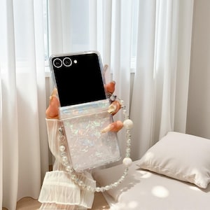 May include: Clear phone case with iridescent glitter and a pearl wrist strap. The case is designed to fit a flip phone and has a clear, protective design. The wrist strap is made of small and large pearls.