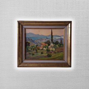 May include: An antique landscape painting in a gold frame. The artwork depicts a coastal scene with a village, trees, and a body of water under a light blue sky. The frame has a gold finish with a white inner border.