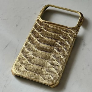 May include: A gold-colored phone case with a textured, scale-like pattern. The case has a raised edge around the camera and a smooth, rounded frame. The overall design is sleek and modern.