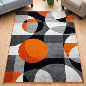 May include: Geometric area rug with a modern design. The rug features large orange, black, gray, and white shapes. The rug has an orange border and is placed on a hardwood floor.