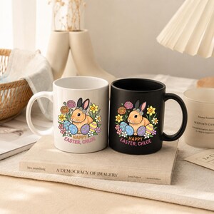 May include: Two ceramic mugs, one white and one black, each featuring a graphic of a brown rabbit surrounded by colorful Easter eggs and flowers. Below the illustration, the text reads "HAPPY EASTER, CHLOE" in a bold, light-colored font.