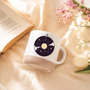 Puede incluir: Una taza de cerámica blanca con acabado moteado y base color canela. El frente presenta un gráfico circular de un disco de vinilo morado oscuro flotando en el espacio, decorado con estrellas, planetas y un pequeño astronauta en el borde.