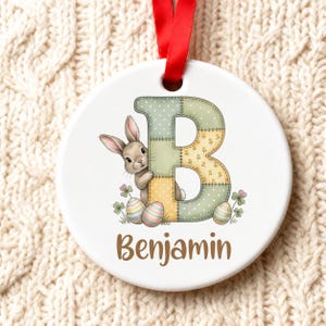Op de afbeelding: Een rond wit keramieken ornament met een grote letter B in een patchworkpatroon in zachte groen- en geeltinten. Een klein bruin konijntje staat naast de letter, omringd door klaver en drie gestreepte paaseieren. De naam Benjamin staat eronder in bruin schrift en aan de bovenkant is een rood lint bevestigd.