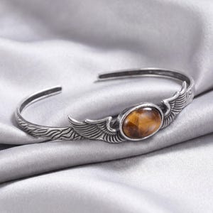 Può includere: Bracciale a polsino argentato con una pietra occhio di tigre ovale al centro. Il bracciale presenta dettagli a forma di ali su entrambi i lati della pietra e un motivo strutturato vicino all'apertura. Presentato su un fondo di raso argentato.