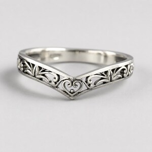 May include: A silver-coloured chevron-shaped ring featuring an intricate openwork design with scrolling filigree patterns and a central heart-shaped motif. The metal has a polished finish with darkened recessed areas to highlight the detailed metalwork.