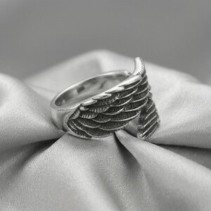 May include: A silver-colored ring with a wing design. The ring features detailed feather textures, with the wing extending around the band. The ring is set against a soft, gray background.