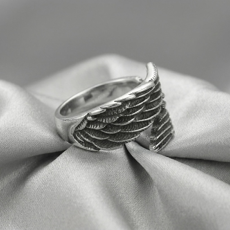 May include: A silver-colored ring with a wing design. The ring features detailed feather textures, with the wing extending around the band. The ring is set against a soft, gray background.