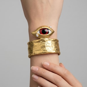 Può includere: Bracciale a polsino dorato con una fascia strutturata e un ornamento a forma di occhio. L'occhio ha un'iride rossa, una sclera bianca e un accento a forma di lacrima. Due piccole pietre trasparenti sono incastonate vicino all'occhio.