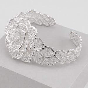 Può includere: Un bracciale a fascia in argento caratterizzato da un intricato design in filigrana. Il fulcro è un grande fiore a più strati con una delicata lavorazione in metallo simile al pizzo. Il bracciale prosegue con foglie stilizzate.