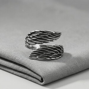May include: A silver-colored ring with an open design, featuring detailed wing-shaped elements. The ring is crafted with intricate feather detailing, creating a textured appearance. The ring is displayed on a gray fabric surface.