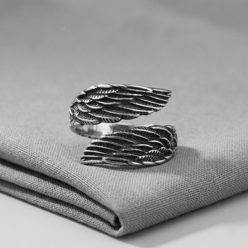 May include: A silver-colored ring with an open design, featuring detailed wing-shaped elements. The ring is crafted with intricate feather detailing, creating a textured appearance. The ring is displayed on a gray fabric surface.