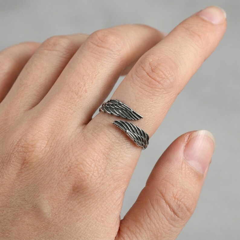 May include: A silver-toned ring in the shape of angel wings. The ring wraps around the finger with the wings extending outwards. The wings have detailed feather textures. The ring is worn on a finger.