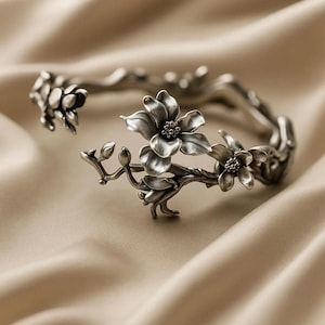 Könnte beinhalten: Ein silberfarbenes Blumen-Armreif-Armband. Das Armband hat detaillierte Blumen- und Astdesigns. Die Blumen haben strukturierte Blütenblätter und kleine, runde Zentren. Das Armband ist an einem Ende offen und liegt auf einer beigen Satin-Oberfläche.