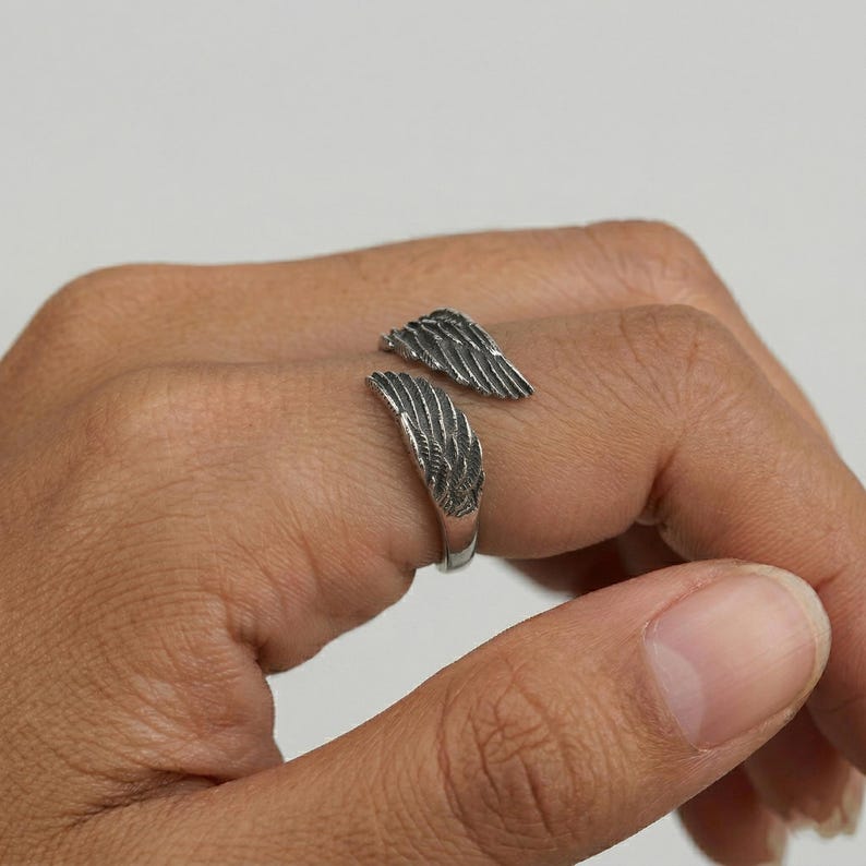 May include: A silver-colored ring with a wing design. The ring features two detailed wing shapes that wrap around the finger. The wings are textured with feather-like patterns, creating a realistic appearance. The ring is worn on a hand.