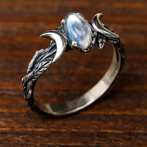 Puede incluir: Anillo de plata con una piedra preciosa ovalada e iridiscente. El diseño celestial incluye medias lunas y detalles de hojas. La banda es lisa y pulida. El estilo es ornamentado y místico. El anillo es probablemente de plata.