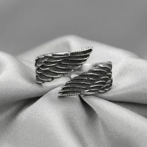 May include: A silver-toned ring featuring a detailed wing design. The ring is open, with each wing forming a separate side. The wings are textured with individual feather details, creating a realistic appearance. The ring is set against a soft, silvery background.