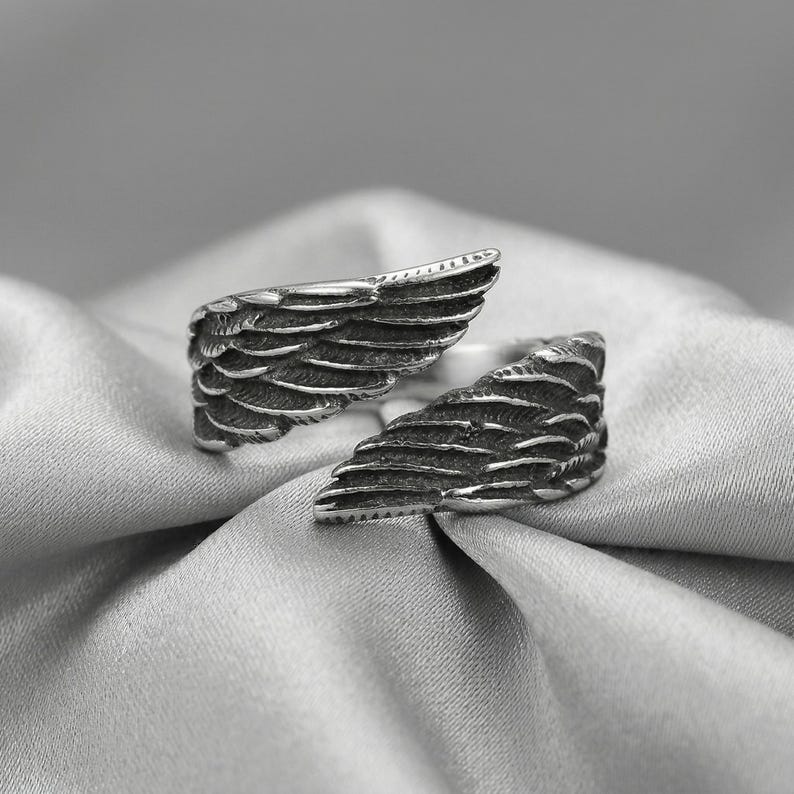 May include: A silver-toned ring featuring a detailed wing design. The ring is open, with each wing forming a separate side. The wings are textured with individual feather details, creating a realistic appearance. The ring is set against a soft, silvery background.